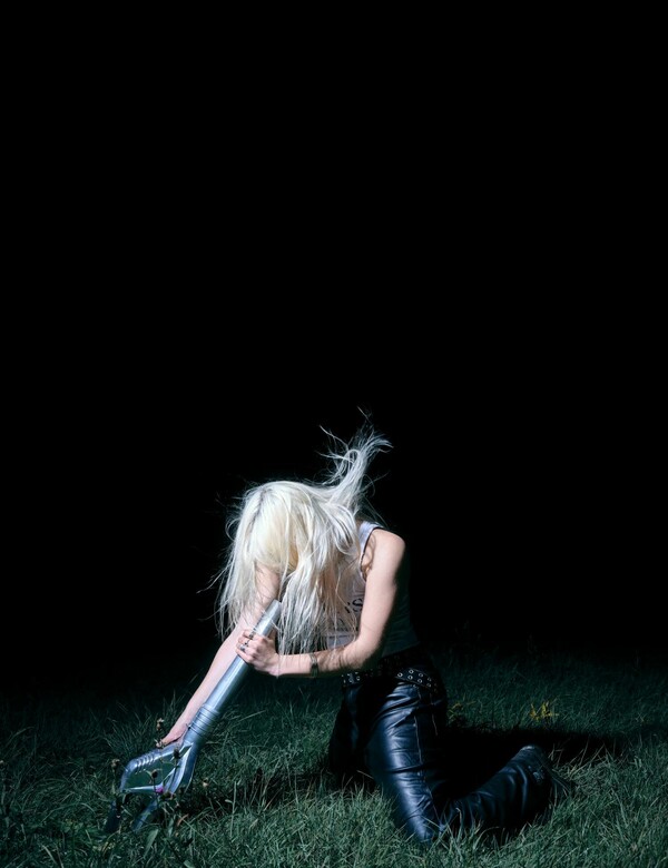 A person with long platinum blonde hair covering their face, wearing a white tank top and leather trousers, kneels on grass while holding a leaf blower aimed at their face, setting their hair into motion.