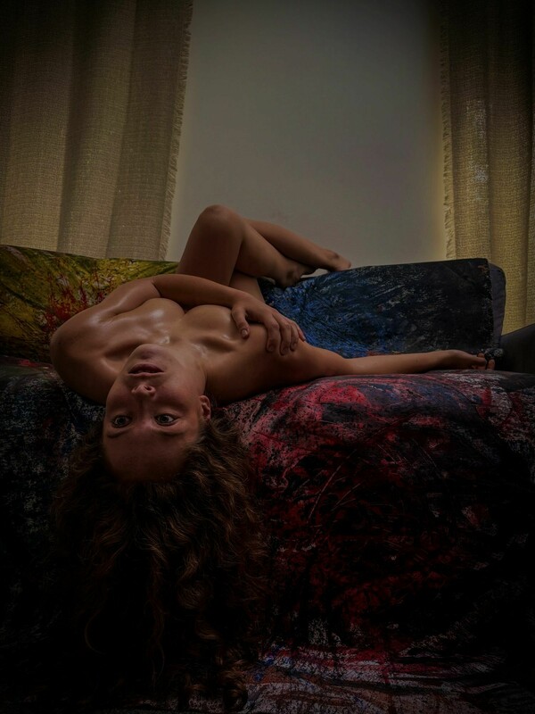 A nude woman lying on a sofa draped with a painted canvas, captured upside down, holding an intense gaze forward.