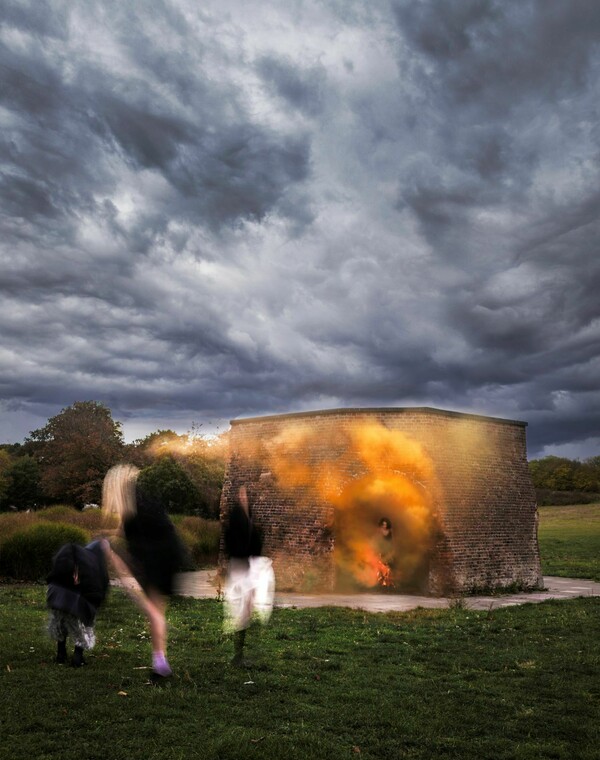 In a setting featuring a grassy field with trees, a red brick building, and a cloudy sky, there are four blurred figures, three gathered outside, and one emerging from the building accompanied by orange smoke.