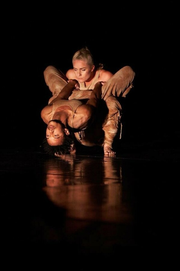 Against a dark background, two figures are illuminated by yellow light as one person squats holding another who is upside down, leaning backward and wrapping their thighs around the squatting person.