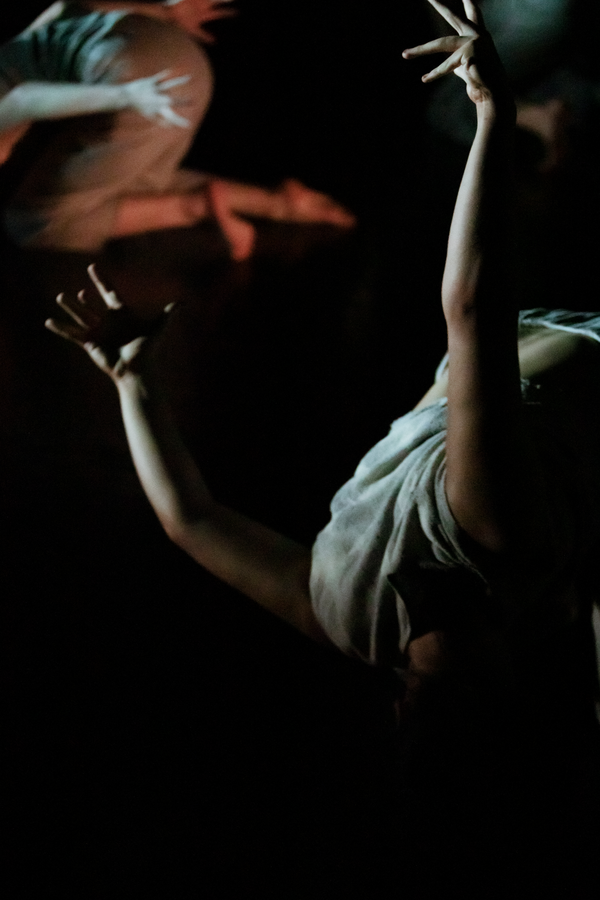 Close-up of a person’s arms with fingers spread, reaching behind their back while lying face down in a lightly lit dark setting.