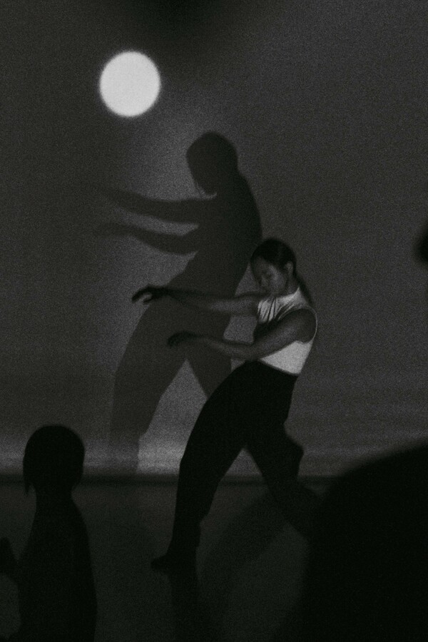Black and white image of three people on stage: two in silhouette – one a close-up profile, the other seated in the distance – and one visible figure wearing a white top and black trousers, posed sideways, leaning back with arms held in front of her body. A backdrop features a round light resembling a full moon.