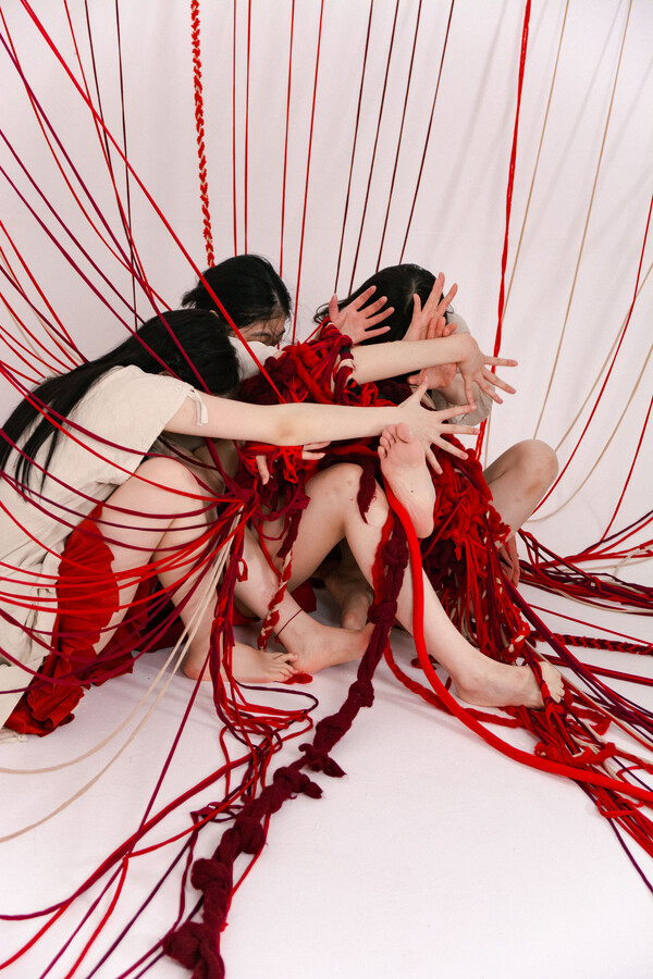 Three people sitting, wearing white tops and short red skirts, stretching their legs and arms forward with fingers spread, caught amid a network of red and white strings cascading from above.