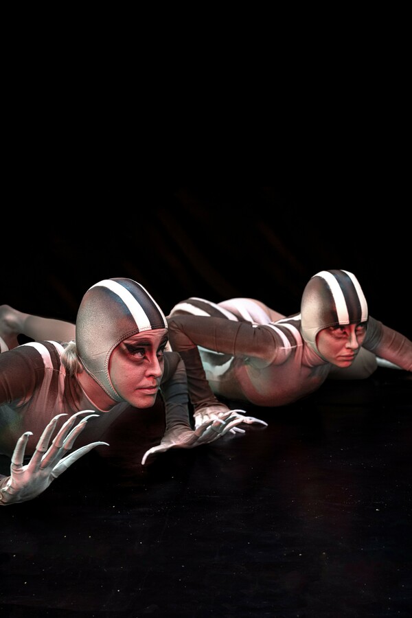 Two people caught in a prowling motion in a dark theatre setting, wearing skintight grey-and-white costumes with matching caps, makeup, and gloves tipped with long, spiky fingers, evoking lizard-like creatures.