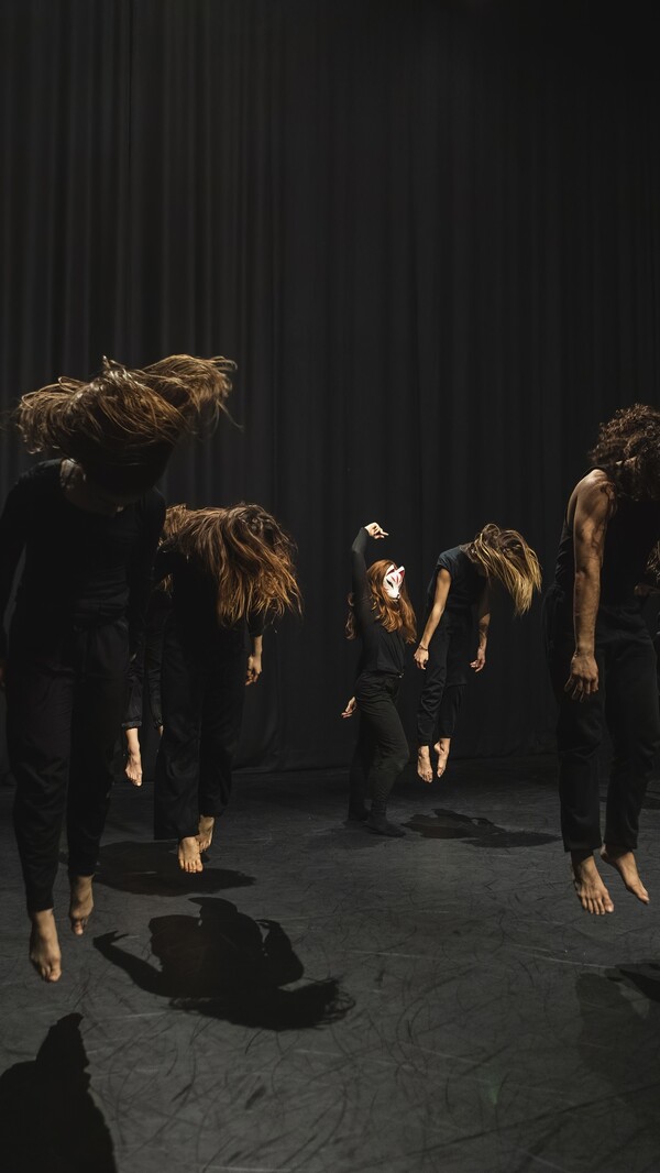 A group of people dressed in full black jumping and looking down, their loose hair set in motion, while one person at the back wearing a white animal mask poses with their right arm raised overhead.