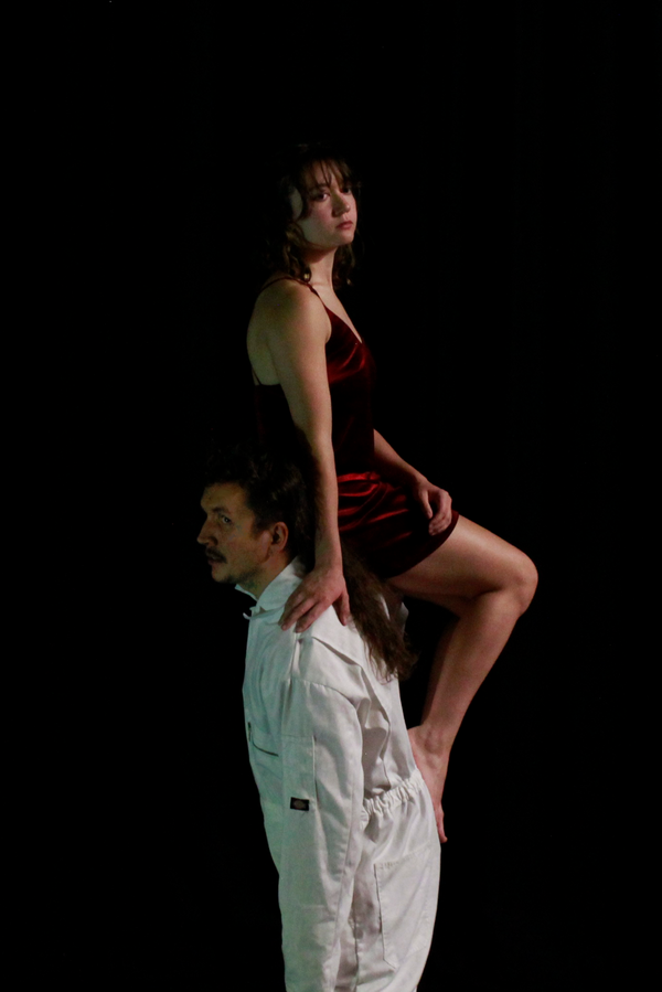 Against a black background, a person in a white overall faces right, holding on their shoulders a person in a short red dress facing left, both gazing ahead.