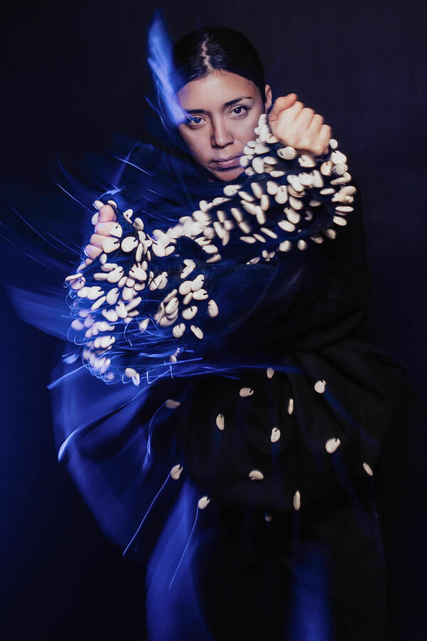 Person in a dark costume embellished with white ornaments, captured mid-motion, crossing their arms at chest-level, gazing forward and surrounded by a blue halo.