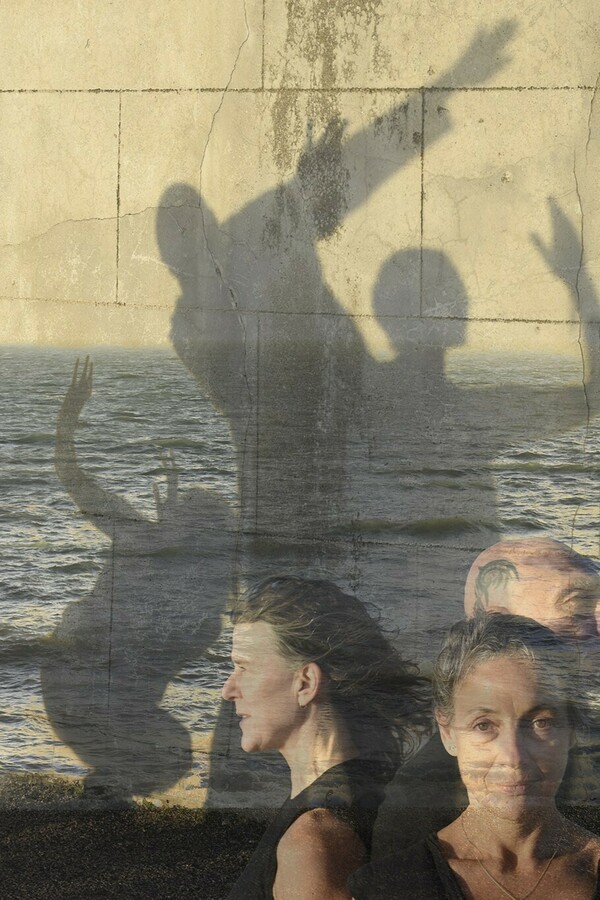 A layered image featuring a sea with two distant figures standing before it, a light brick wall casting shadows of three posing figures, and three people shown from the shoulders up, one facing forward, with two behind her in profile facing right and left.