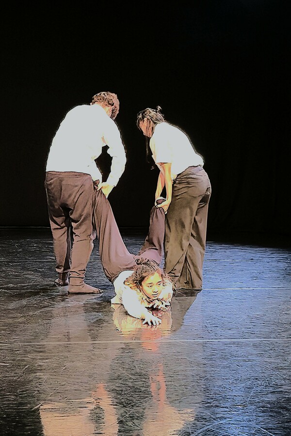 Three people, all wearing white t-shirts and black trousers, are in an all-black theatre setting, with two standing back to the audience, lifting the legs of a third person lying on their stomach, looking forward and reaching ahead.