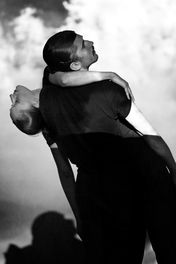 Black-and-white photo of two people, one in a black T-shirt and trousers leaning back and looking sideways while holding another partly hidden behind them with an arm wrapped around their neck.