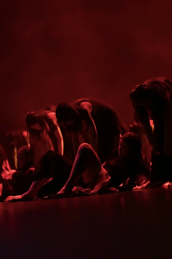 In red light, two rows of people, with the front row lying on their backs propped on elbows with knees bent, and the row behind leaning forward with arms hanging down.