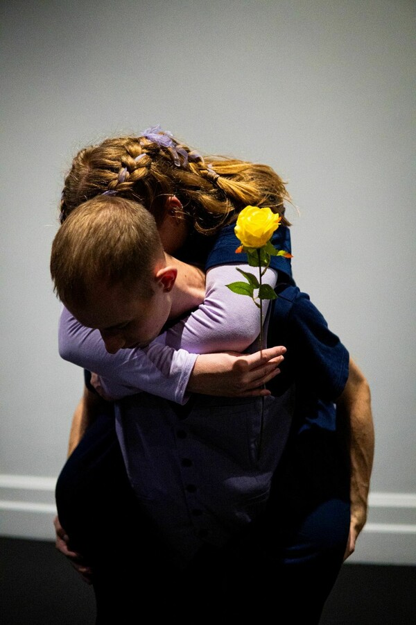 A person with short hair wearing a blue shirt leans forward with their face down, holding on their back another person whose hair is long and partially braided, wearing a navy T-shirt layered over a light purple long sleeve, and holding a yellow rose in their hand.