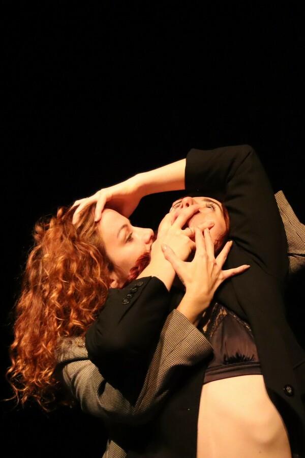 A person dressed in a satin bra and black blazer leans backward, covering their open mouth with one hand and extending the other arm above their head to touch the head of another person embracing them from behind, who is wearing a grey blazer and has long, curly ginger hair.