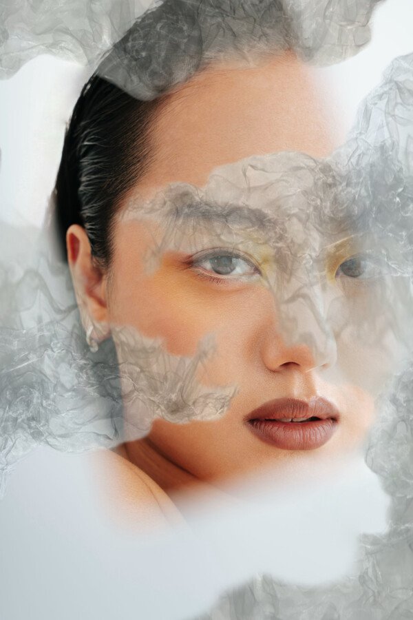 A double-layered image featuring a portrait of a person with slicked-back black hair, yellow eyeshadow, and brown lipstick, looking toward the viewer with their mouth slightly open, overlaid with white and grey abstract shapes resembling clouds.