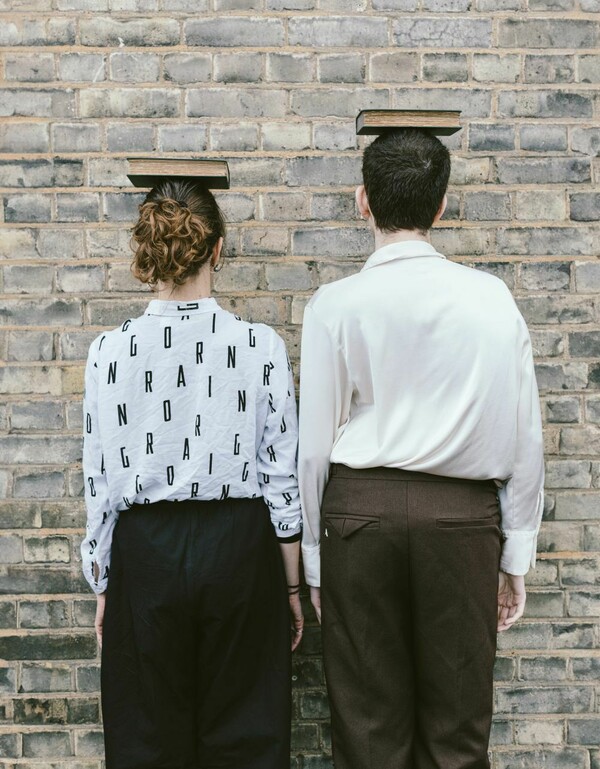 Two people wearing white shirts – one patterned with letters – and dark trousers stand with their backs to the viewer in front of a brick wall, arms at their sides, each balancing a book on their head.