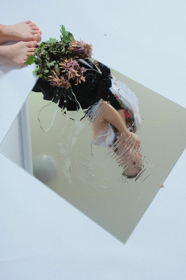 A woman’s smudged reflection in a mirror as she looks at herself, with withered flowers resting in the mirror’s top left corner, while the woman’s feet and the hem of her skirt are visible below.