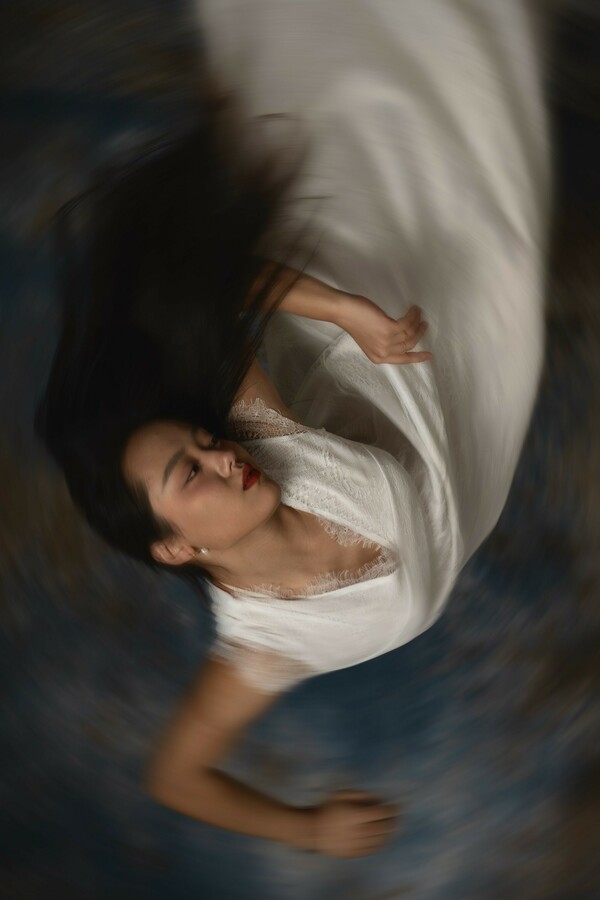 Blurred image of a person with long black hair, seen upside down, wearing a long white dress, leaning sideways with arms raised to shoulder level and elbows bent at right angles.