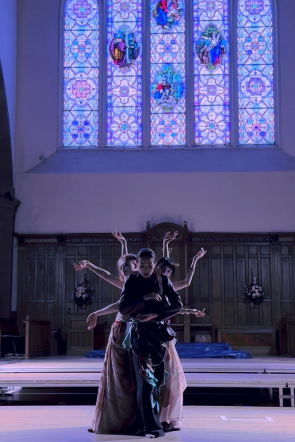 A person in a black costume stands at the front, looking downward, while a group behind her extends their arms to create a geometric pose against a background suggesting a church setting.