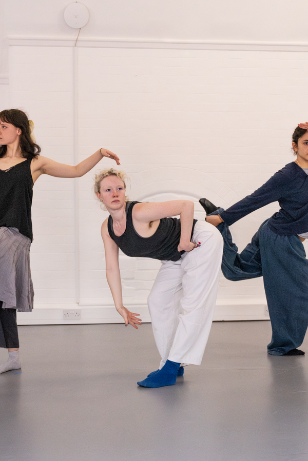Three people pose in a line before a white wall: one lifts a leg forward with arms outstretched, another bends sharply forward reaching down, and the third holds a leg back with one arm while raising the other as if scanning the distance.