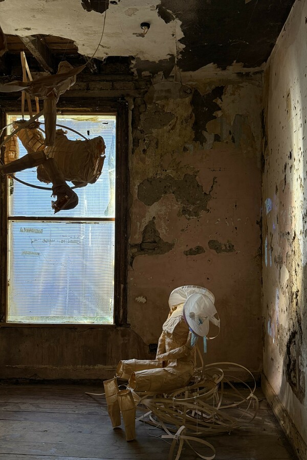 In a decrepit room with peeling paint and a rundown wooden floor, a distorted mannequin sits on an abstract structure by a window, while another abstract structure hangs from the ceiling.