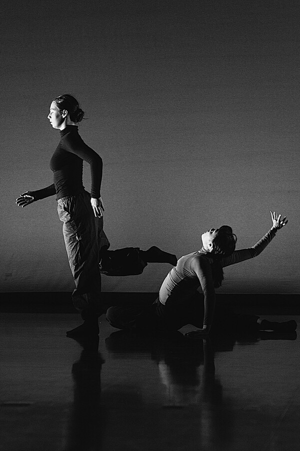 Black and white image of two people, one standing sideways with a leg bent backward, nearly touching the other, who sits with one leg bent and the other extended back, leaning on one arm and reaching the other behind them.