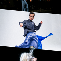 This image is divided into three parts. On the top, woman standing on stage in front of a group of musicians, wearing a long dark skirt and white jacket, captured in between movements with her arms by her sides. In the centre, person wearing a navy shirt and two blue shirts styled as a skirt with red sneakers, caught mid-movement, setting the fabric into motion. On the bottom, a person with long platinum blonde hair covering their face, wearing a white tank top and leather trousers, kneels on grass while holding a leaf blower aimed at their face, setting their hair into motion.
