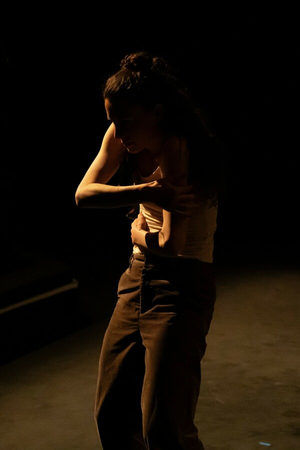 A woman with long hair, wearing a white tank top and dark trousers, is illuminated by low light as she wraps her arms around herself and curves her back.