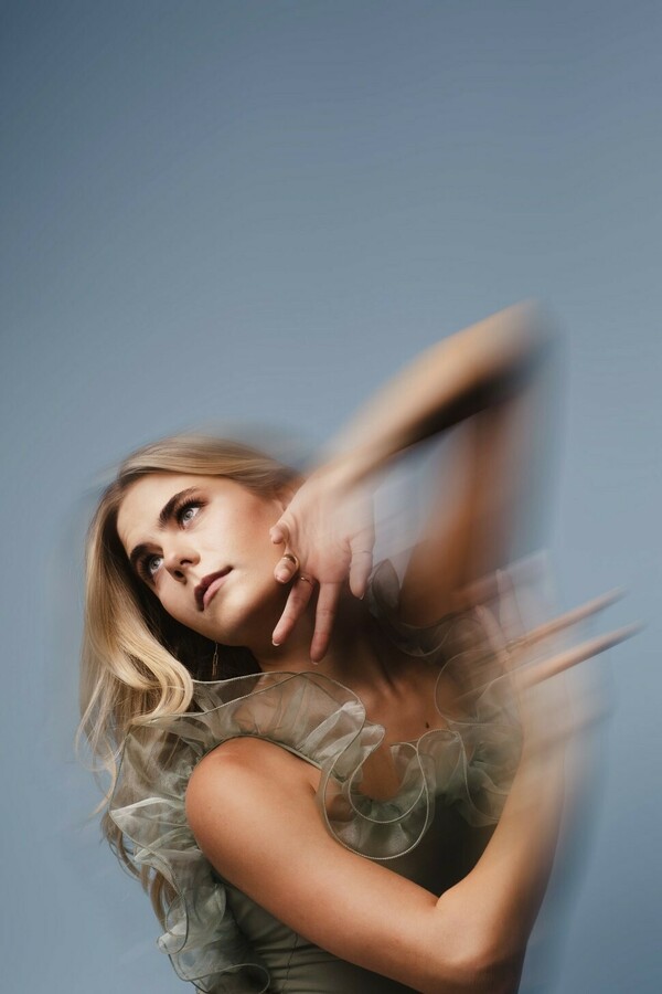 A person in a grey dress with ruffled lining poses with arms raised, blurred from movement, leaning slightly backward and looking sideways against a light grey background.