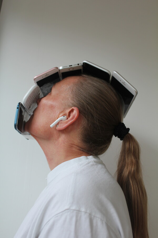 Profile of a person with long grey hair tied in a ponytail, wearing a white T-shirt and white wireless headphones, looking upward with a row of mobile phones atop their face.