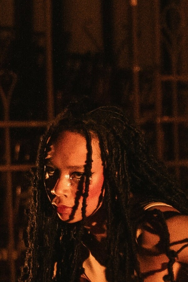 Person with long black braids wearing a nude camisole, shown from the shoulders up at a diagonal angle, holding an intense gaze under warm light.