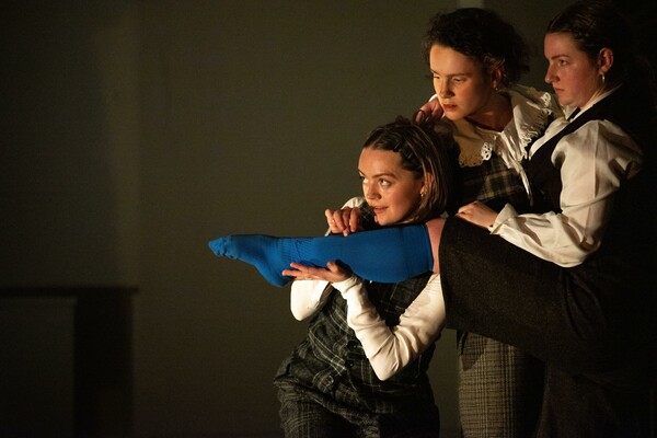 Three people wearing white shirts with dark dresses or sets over them, resembling school uniforms, hold up the leg of one person dressed in a cobalt blue stocking. The leg points to the left of the composition, while the group, clustered on the right, look in the same direction as if scheming or plotting something together.