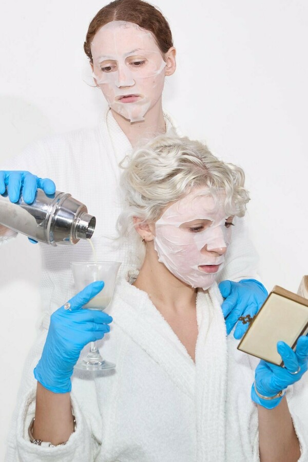 Two people in white bathrobes, blue rubber gloves, and sheet face masks captured from the waist up, one seated lower holding a book and a glass while the other stands behind pouring white liquid from a silver cocktail shaker into the glass.