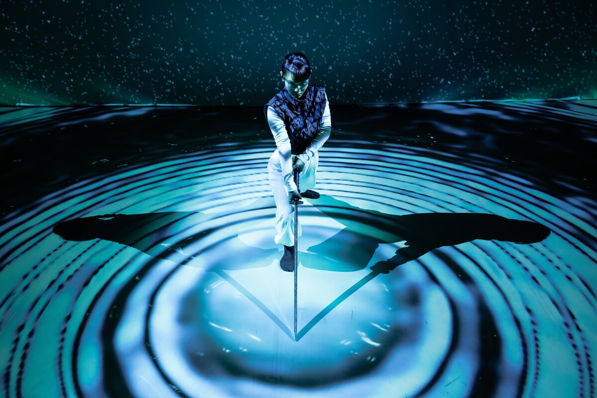 A woman wearing white trousers, a white long-sleeve top, and a navy gilet leans forward onto a long sword, its tip resting at the centre of concentric circles made of blue light and deep shadow. Behind her, a backdrop of scattered light dots against darkness evokes a starry night sky, with a faint green haze resembling northern lights. Her shadow stretches symmetrically to both her left and right, reinforcing the image’s geometric composition. She appears focused and contemplative.