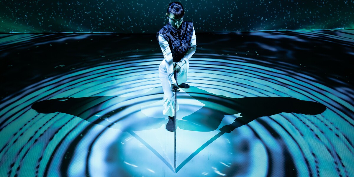 A woman wearing white trousers, a white long-sleeve top, and a navy gilet leans forward onto a long sword, its tip resting at the centre of concentric circles made of blue light and deep shadow. Behind her, a backdrop of scattered light dots against darkness evokes a starry night sky, with a faint green haze resembling northern lights. Her shadow stretches symmetrically to both her left and right, reinforcing the image’s geometric composition. She appears focused and contemplative.