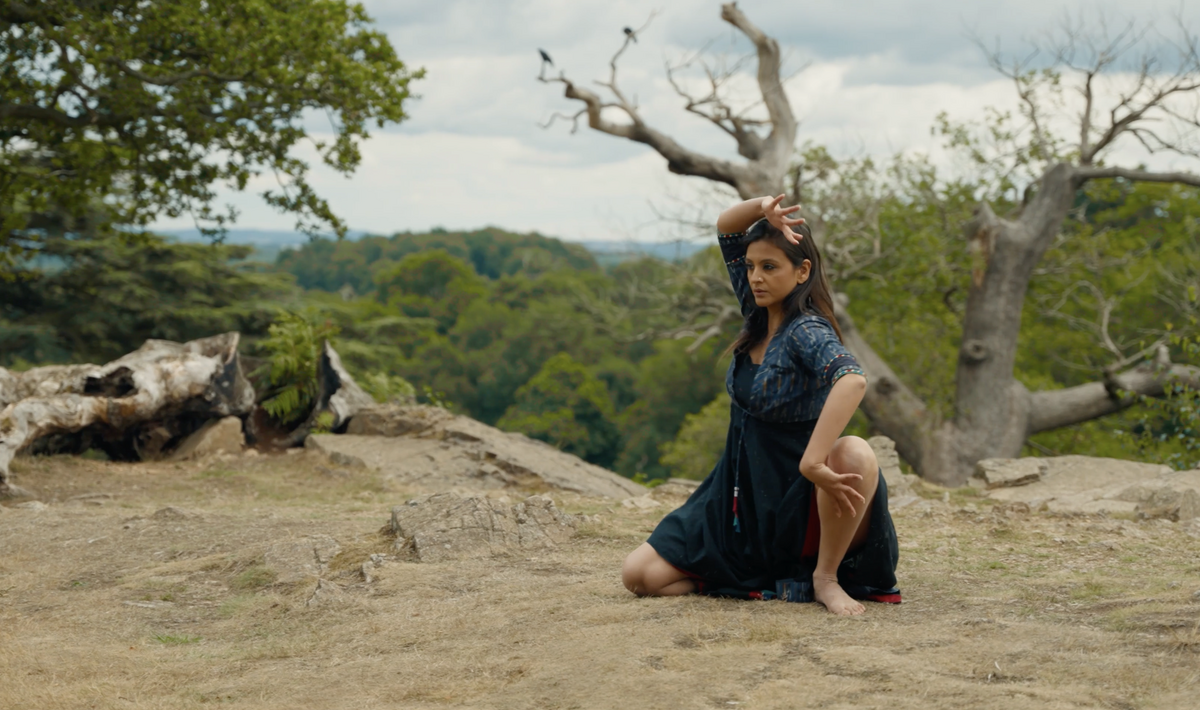 A woman in a dark blue and black dress kneels barefoot on dry, rocky ground, holding a poised classical dance position with one arm curved above her head and the other bent near her knee. Behind her stretches a wide landscape of green hills and trees, under a dramatic sky. A gnarled, leafless tree adds striking contrast in the background.