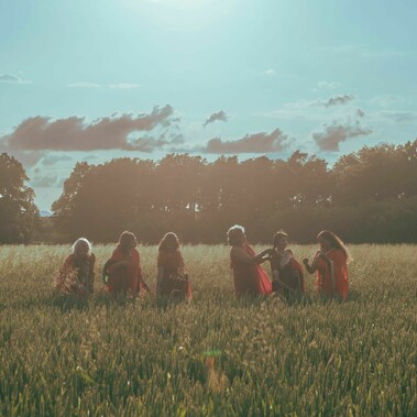 A group of six South Asian women in red and orange saris sit in a field of tall grass at golden hour, lit by the soft glow of the setting sun. They smile and interact warmly with one another, framed by silhouetted trees and a wide, open sky filled with clouds. The image feels intimate, joyful, and grounded in community.