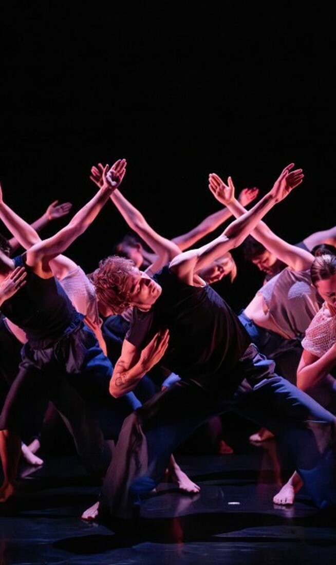 A group of dancers move in sync of stage underneath pink lighting. Their knees are bent, one arm is bent into their chest and the other arm is reaching out.