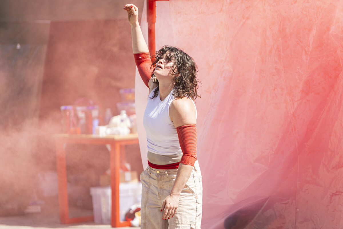 A white woman with black curly hair is stood in front of a red pole. She is surrounded by red smoke. Her arm is stretched above her head and she looks distressed.
