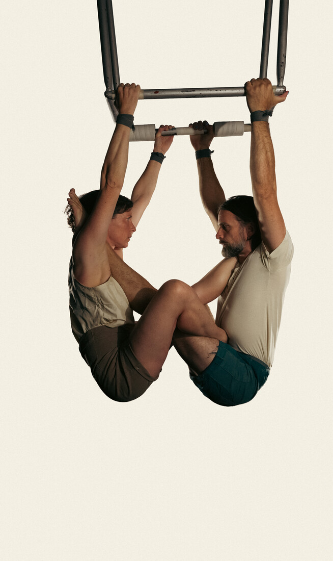 Two people are suspended high in the air from a frame, their legs wrapped around each other as they look into each other's eyes.