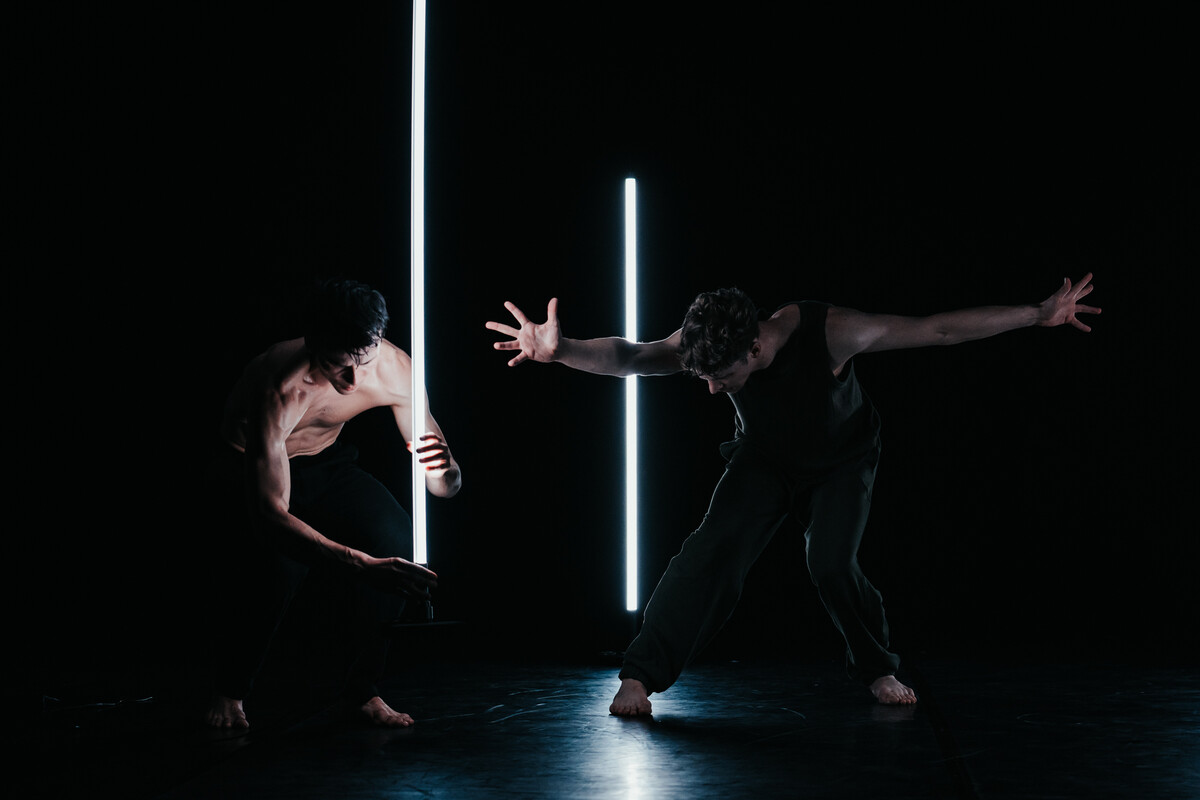 Two male dancers perform a duet with LED light tubes. One crouches near a glowing tube while the other extends his arms wide in a strong stance. Short: Two male dancers perform duet with vertical LED tubes.