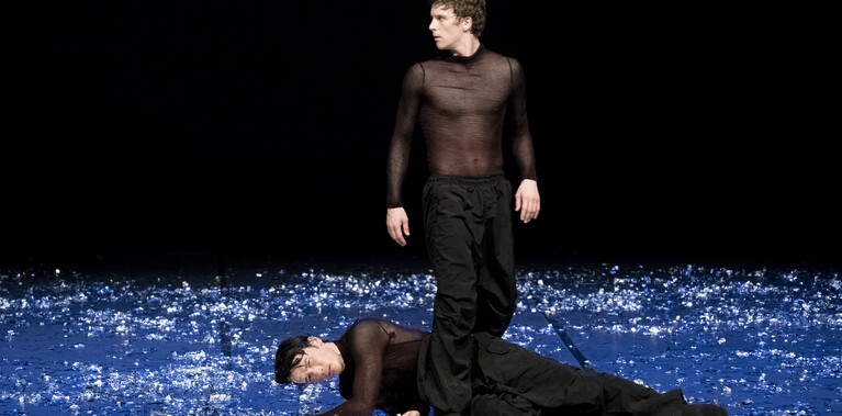 Two male dancers on a stage scattered with reflective fragments. One lies on the ground while the other stands over him, gazing outward into the darkness. Short: Two male dancers; one lies on floor, the other stands over him.