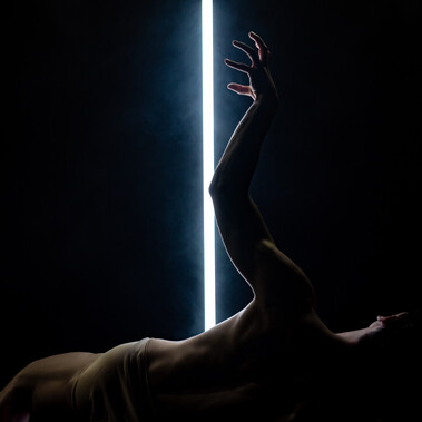 A male dancer lies on the floor, his torso arched back with one arm extended upward. Behind him, a vertical LED light cuts the stage with stark illumination. Short: Male dancer lies back on floor, one arm raised, LED tube behind.