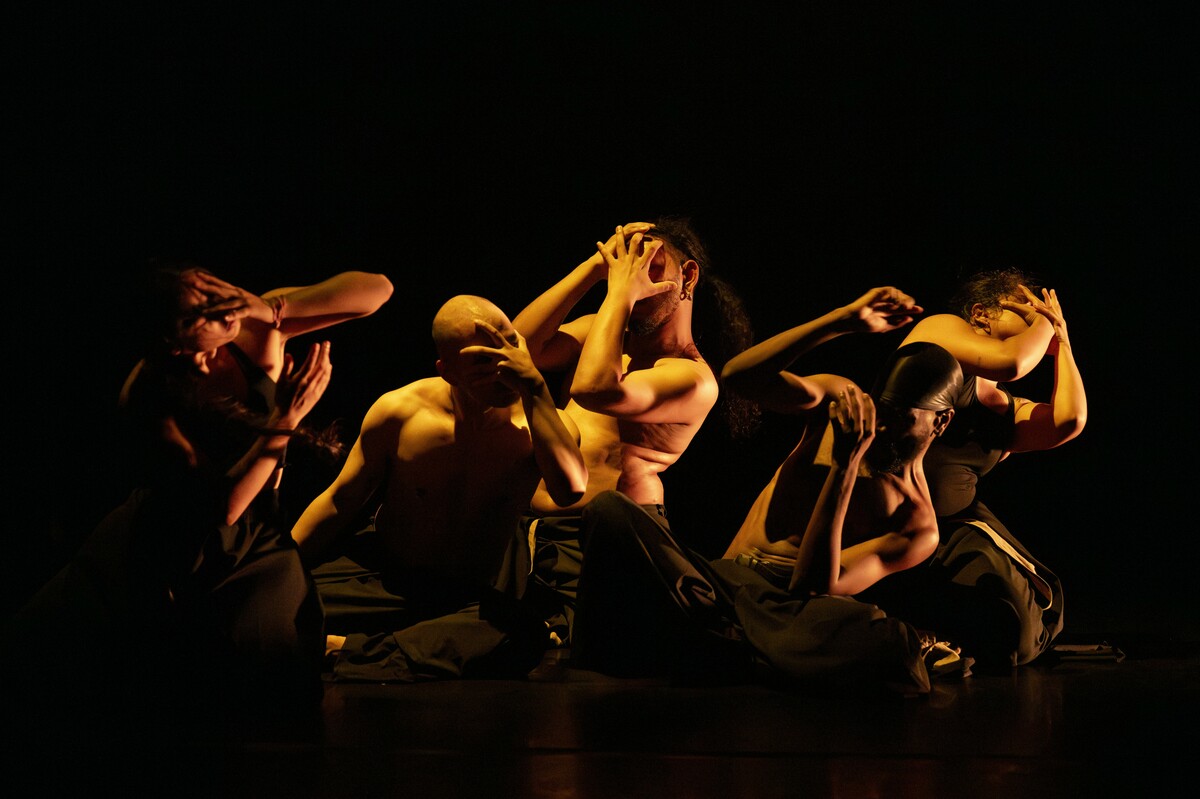 A group of people wearing black trousers—some with black tops, others shirtless—are kneeling and covering their faces with their hands, illuminated by warm light that contrasts with the surrounding darkness.
