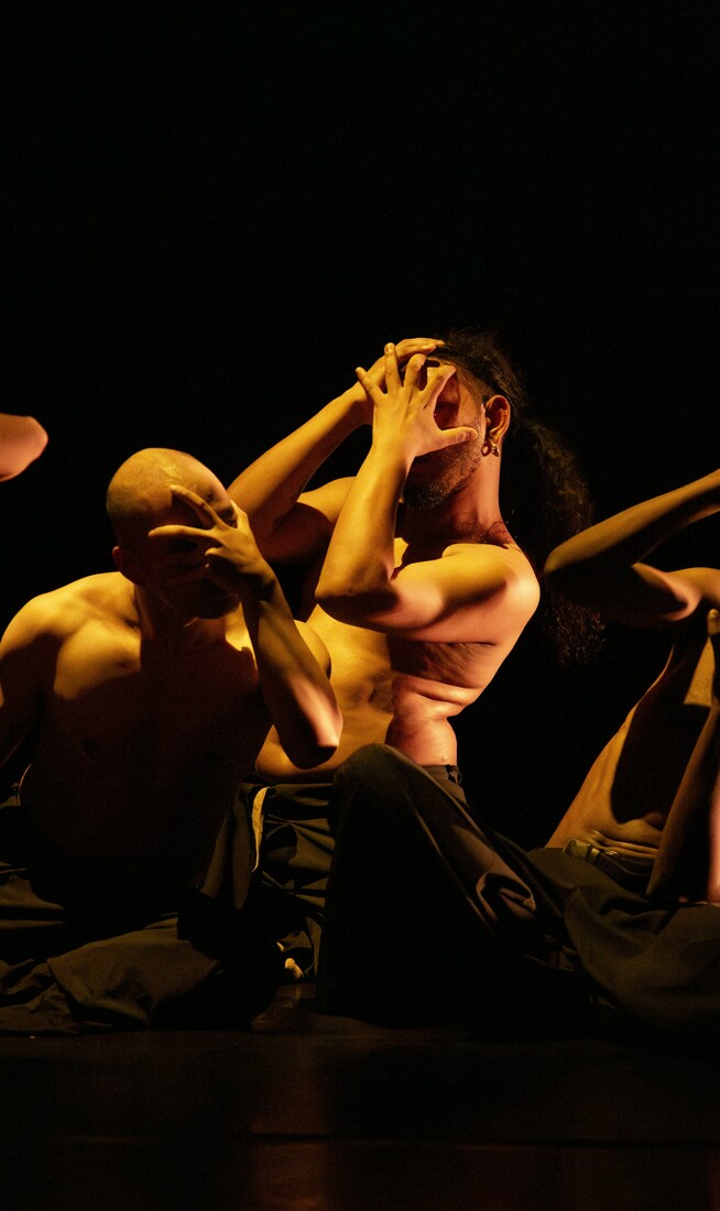 A group of people wearing black trousers—some with black tops, others shirtless—are kneeling and covering their faces with their hands, illuminated by warm light that contrasts with the surrounding darkness.