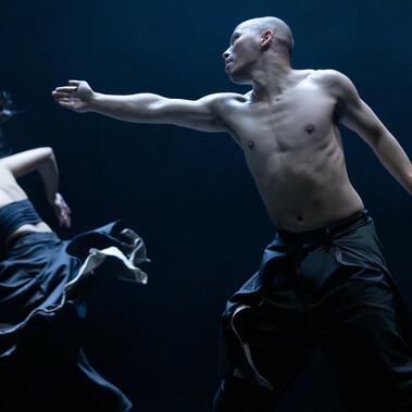 A person wearing black trousers, shirtless and bald, looks to the left and stretches their arm in that direction toward another person in a black top and skirt, caught mid-turn.
