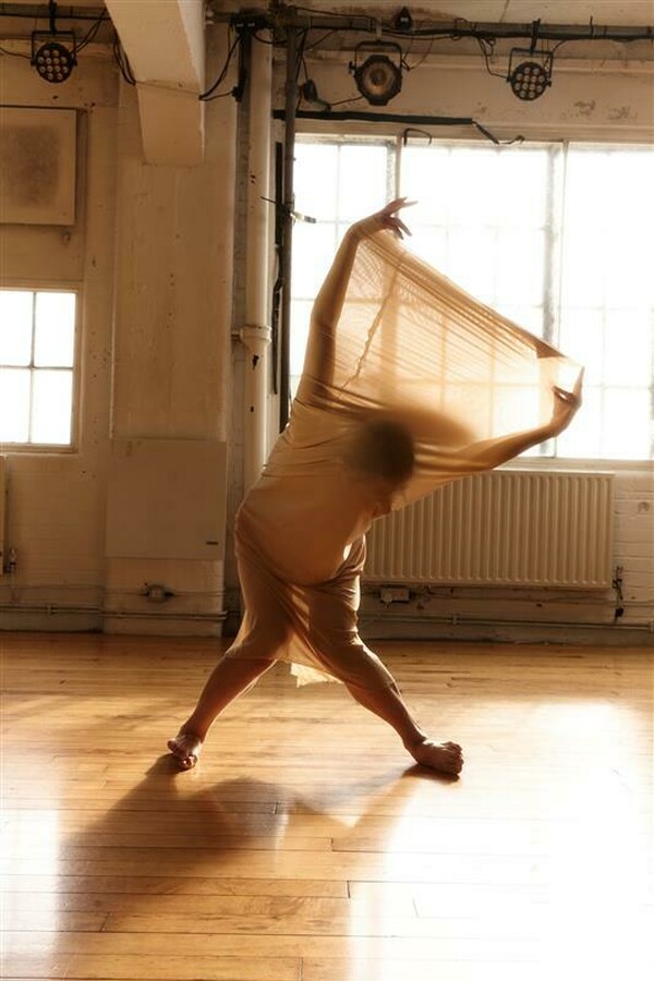 Silhouette of a person standing with legs apart and knees bent inward, leaning forward as they lift their arms straight behind their back in a symmetrical pose, wearing translucent beige fabric that stretches from knees to arms as they extend.