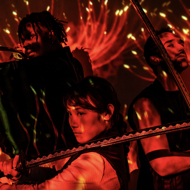 Three people hold swords in different positions: one horizontally across their shoulder, one pointing upward, and one angled outward. They are lit by red light that forms abstract, stylized plant shapes over them and the backdrop.