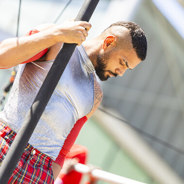Brown-skinned man with a short mohawk and beard, wearing a grey tank top and red checkered trousers, shown from the waist up. He holds a black pole with one hand and looks away from it, gazing downward.