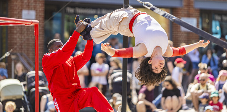 A Black man wearing a red tracksuit is holding the feet of a white woman suspended backwards on a black pole. She is wearing a white top and beige trousers, and her arms are outstretched.