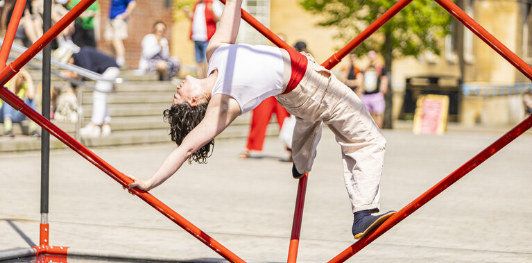 White woman with mid-length curly brown hair, wearing a white tank top and beige trousers, is suspended within a red cube-shaped sculpture made of rods, positioned diagonally on one edge. She grips two edges with her feet, leans back, holds one edge with one hand, and reaches toward the top edge with the other.